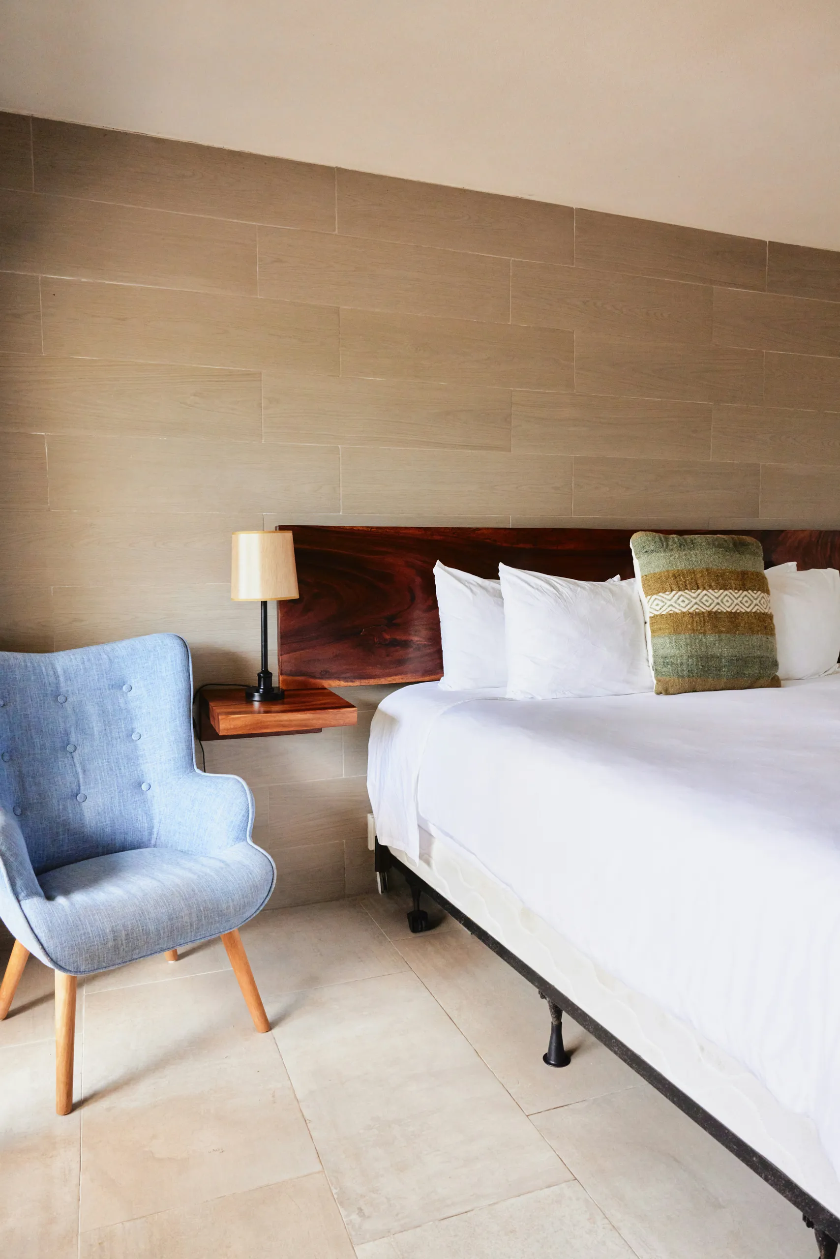 Habitación con cabecero de madera, ropa de cama blanca y un sillón azul junto a la cama.